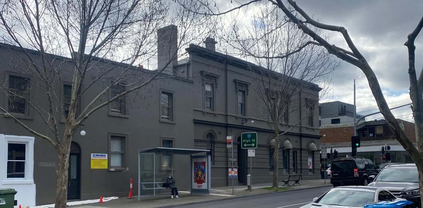 A High Street Armadale commercial building in its original dark charcoal render before repainting, with drop sheets laid along the footpath and a bus shelter against the frontage