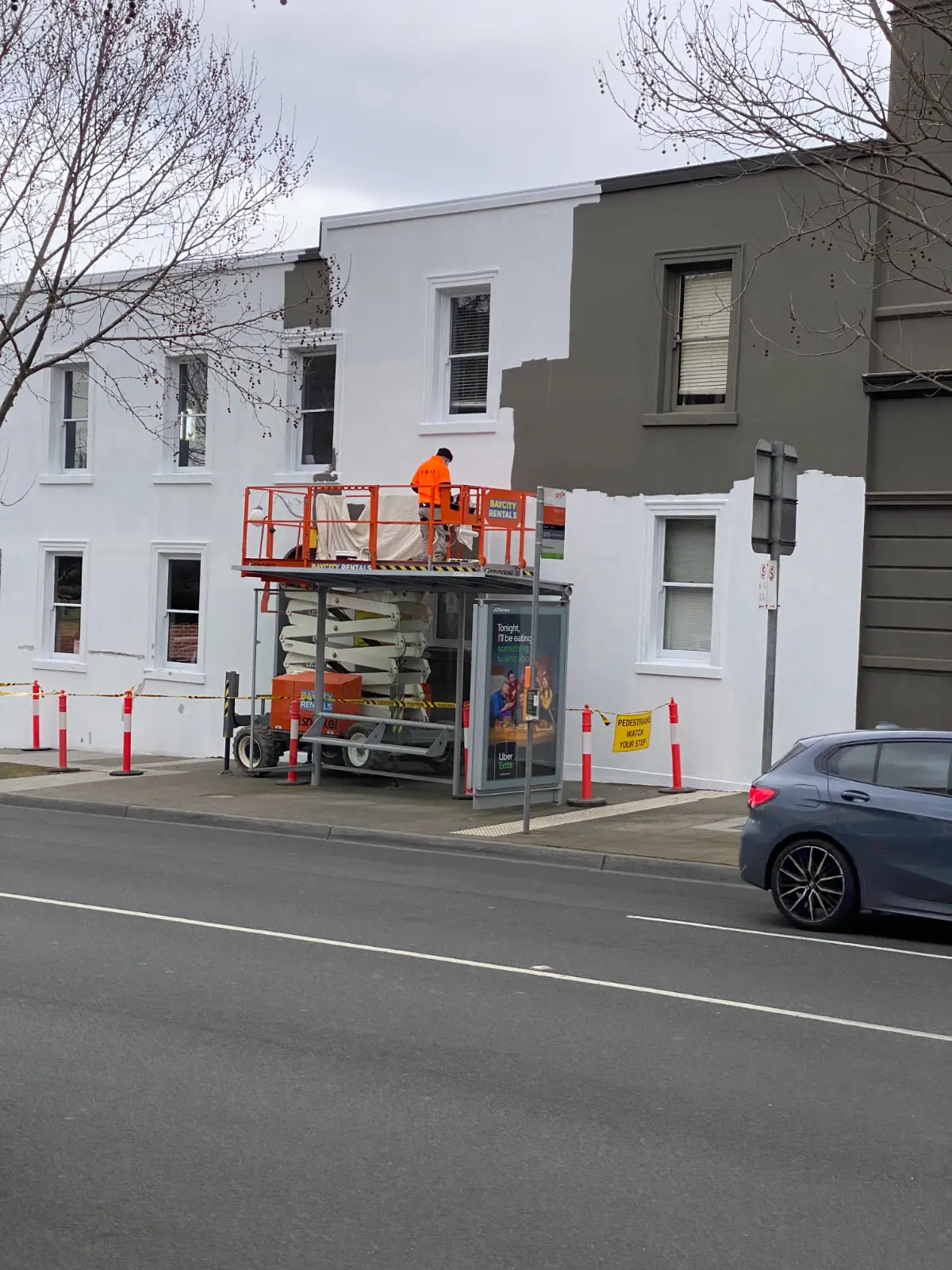 A Jetblack Painting painter on a scissor lift repainting a rendered commercial facade in Armadale, white meeting the original charcoal on a hard edge, with cones and bunting around a live bus stop
