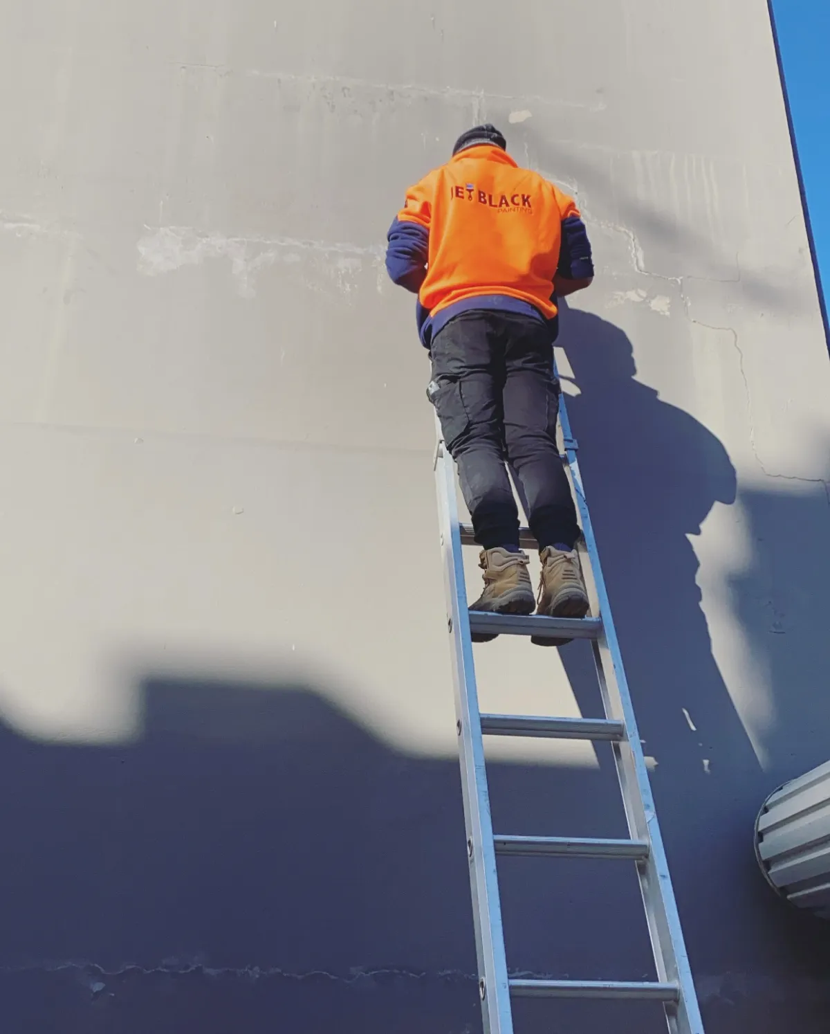 A Jetblack Painting painter in branded orange workwear on an extension ladder, working the upper cream section of a two-tone commercial wall in Armadale above a dark navy band