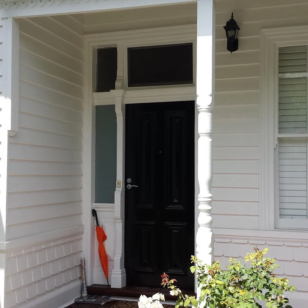 Finished entry of a period weatherboard home in Armadale, off-white boards and shingle course with a white turned verandah post, black front door and black coach light