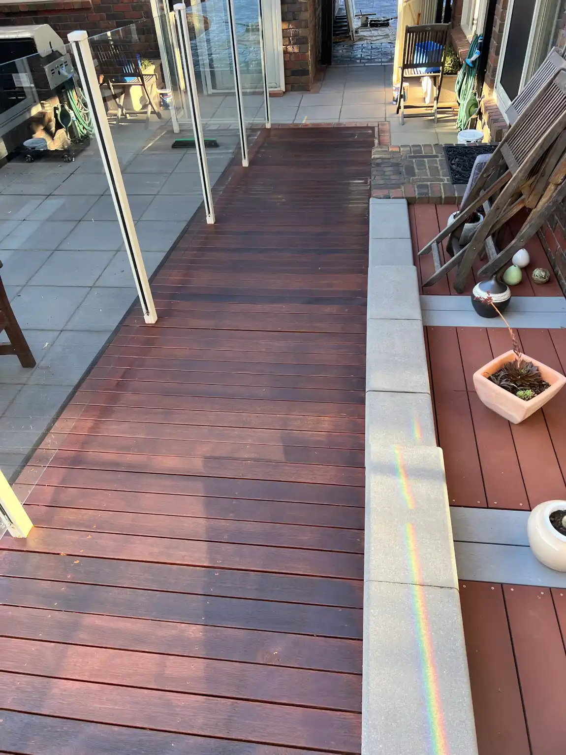 Hardwood deck along the side of a Aspendale Gardens home finished in a deep red-brown, running beside glass pool fencing and a paved courtyard