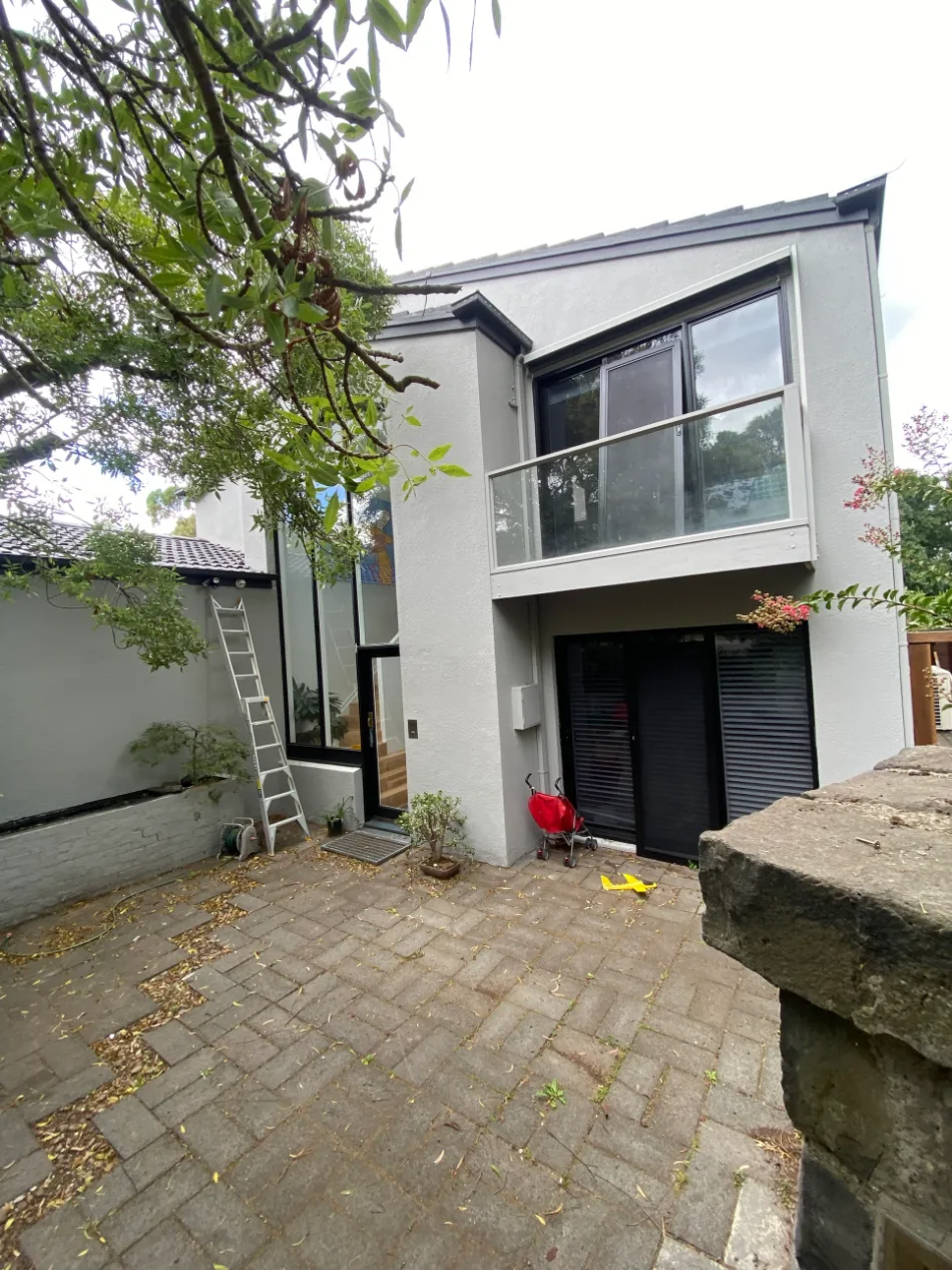 Two-storey rendered home in Balwyn after exterior painting, with light grey render walls, a glass-fronted balcony and black window frames over a paved courtyard