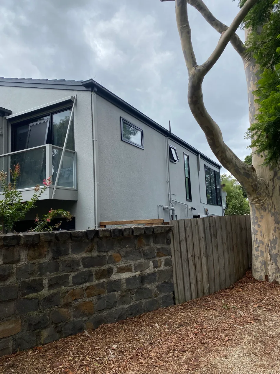 Long side elevation of a rendered two-storey home in Balwyn after exterior painting, light grey walls and downpipes above a bluestone wall