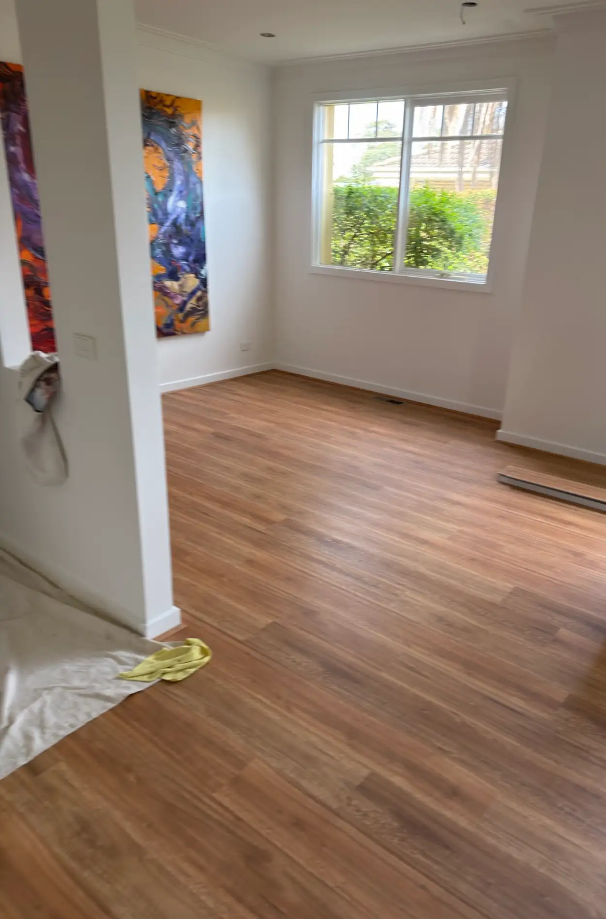Living room in a Beaumaris home after repainting, with white walls and ceiling behind two tall abstract artworks, a board floor and a window looking onto a hedged garden