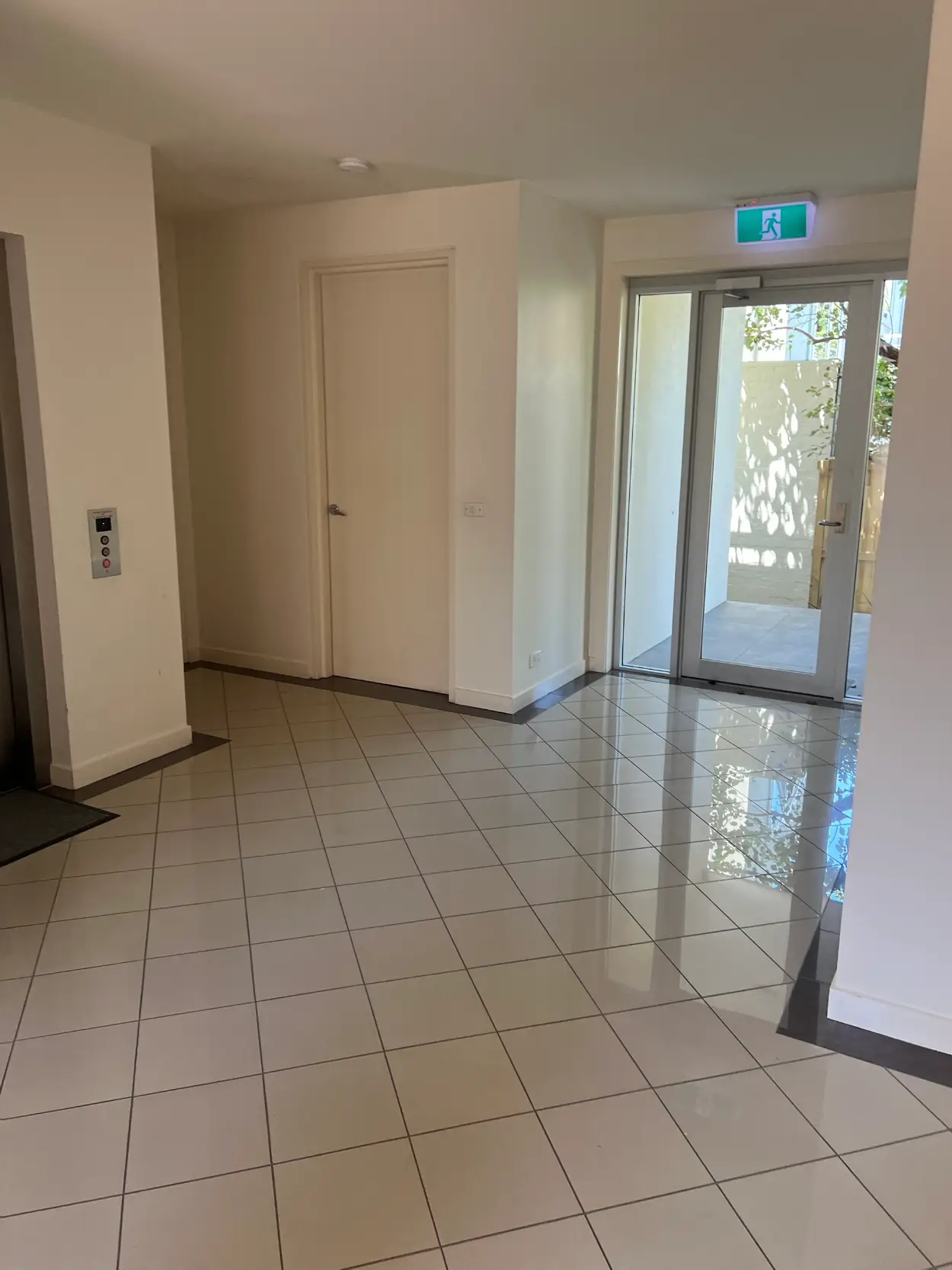 Ground-floor lift lobby in a Beaumaris apartment block after repainting, with white walls above a tiled floor, a lift call panel and a glazed entry door to the street