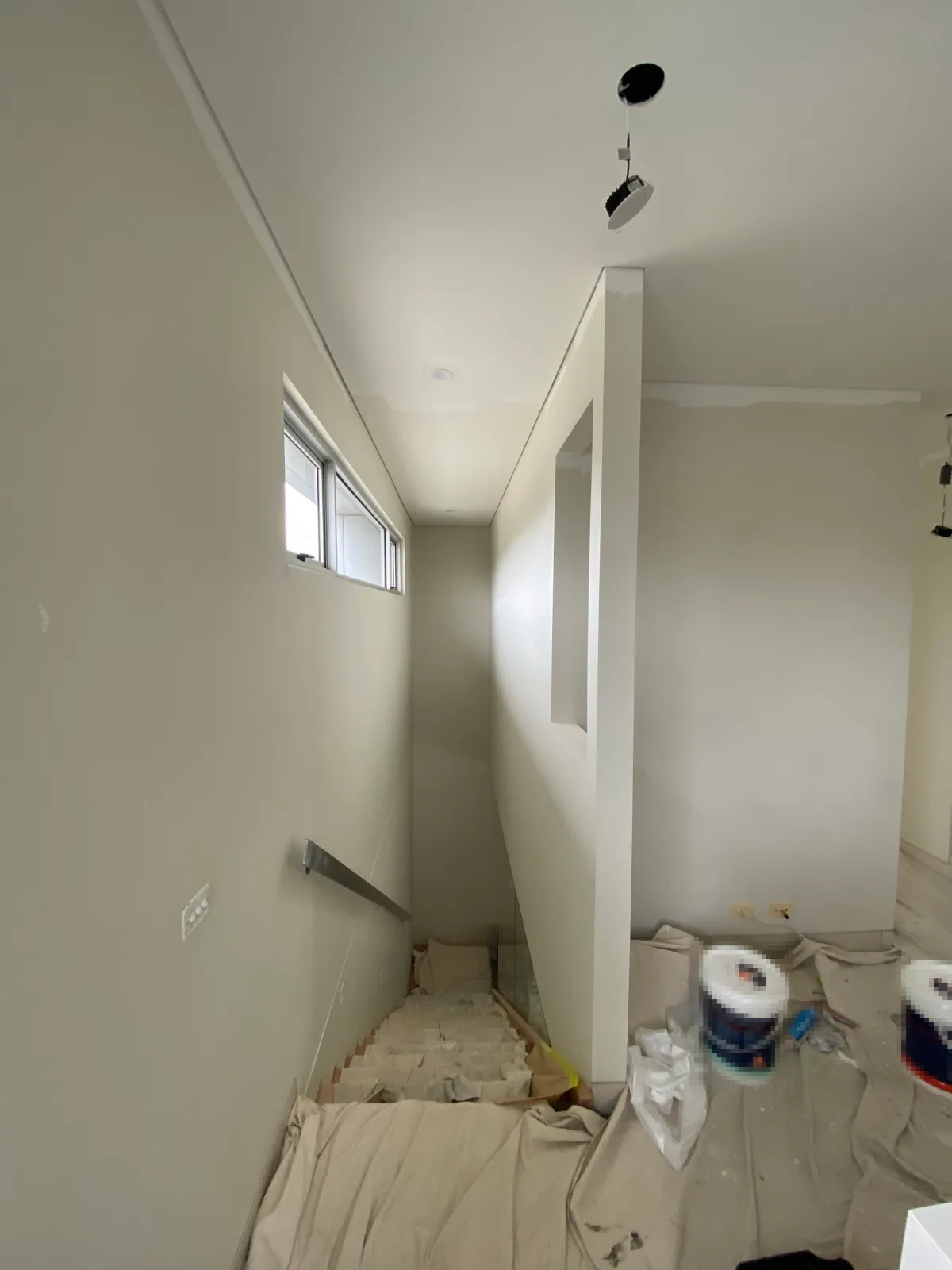 Stairwell of a Beaumaris home mid-repaint, walls and ceiling in white with a high window and an internal wall opening, the stairs and floor under drop sheets and paint tins on the landing