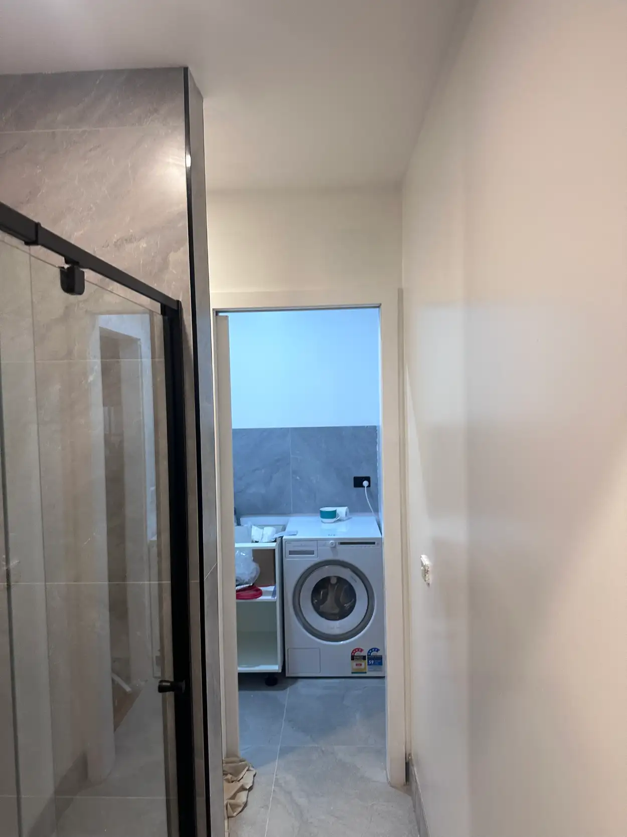 Renovated Bentleigh bathroom looking through to the laundry, with white walls, ceiling and door frame beside a grey stone-look tiled shower wall and black-framed screen