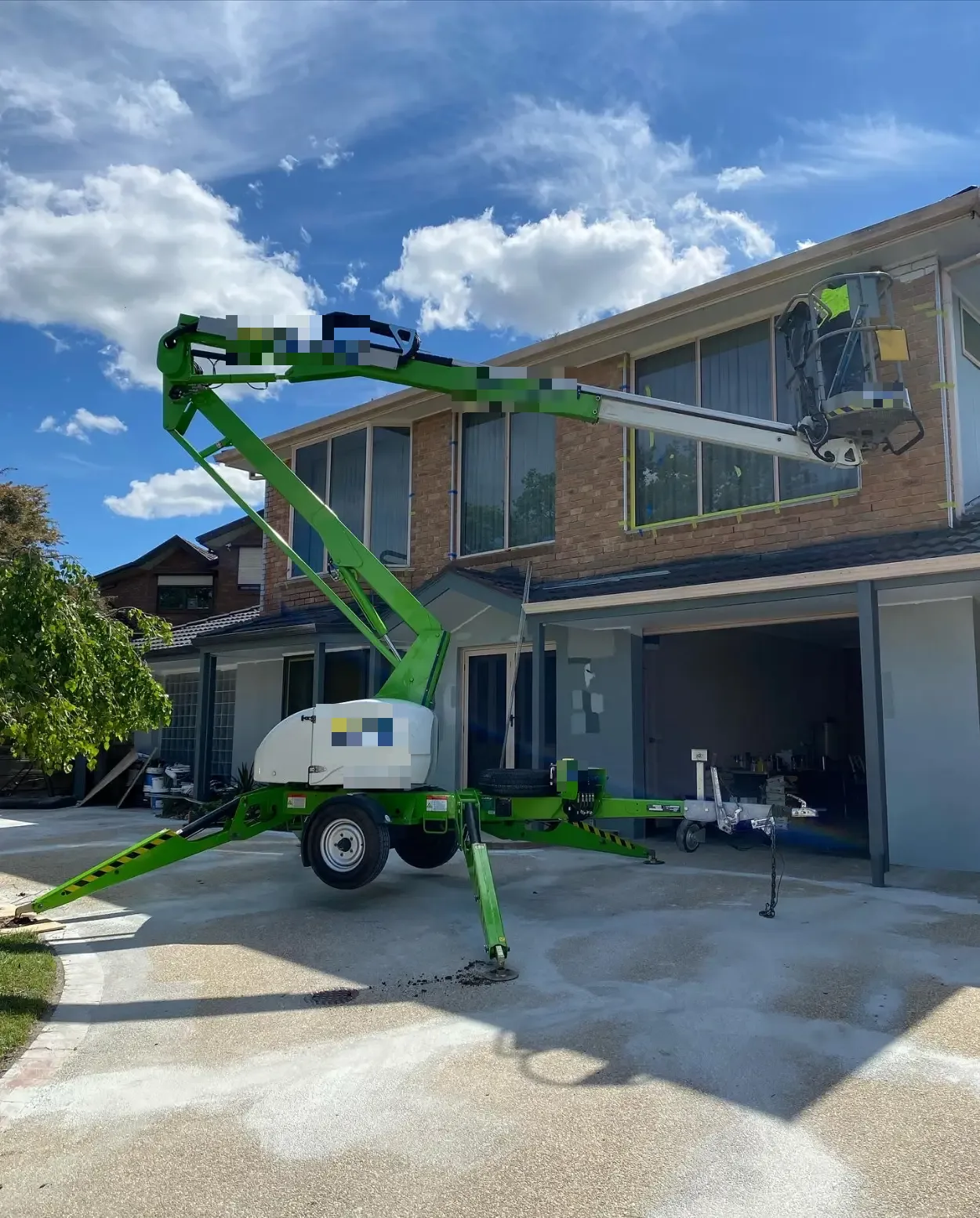 Trailer-mounted boom lift set up on the driveway of a two-storey Berwick home, its platform raised to the upper-level brickwork with the windows masked off ready for painting