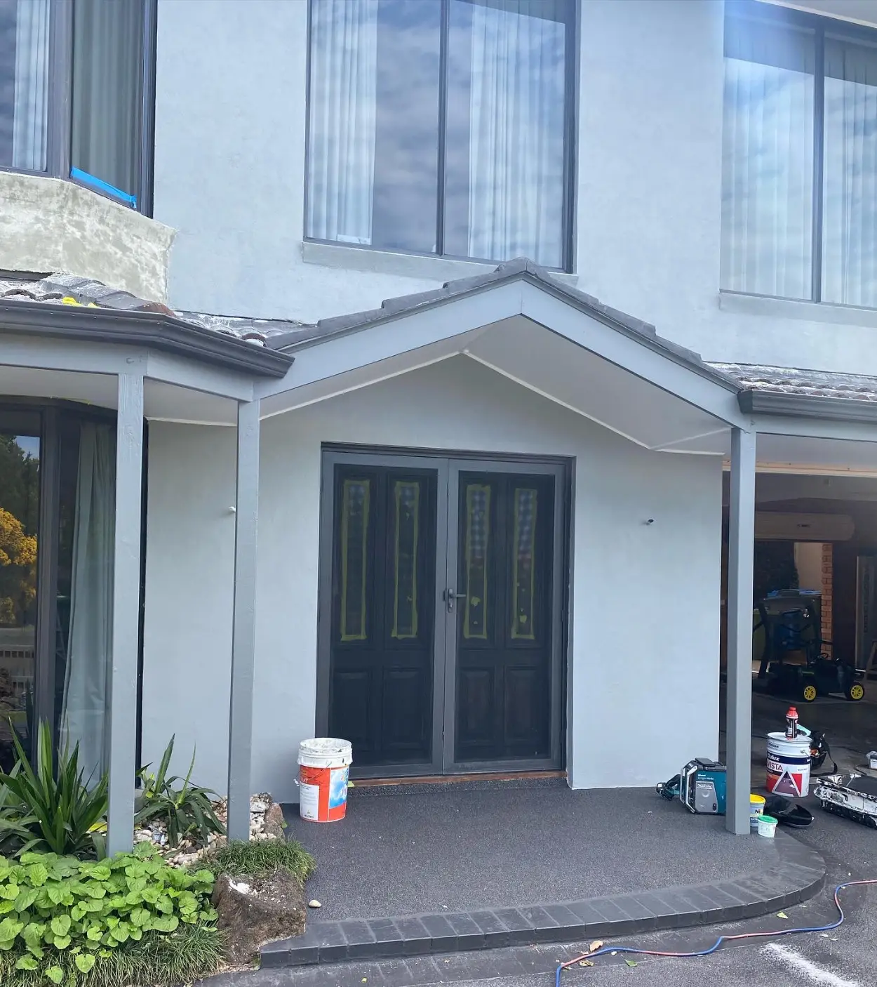 Entry portico of a Berwick home during an exterior repaint, with pale grey rendered walls, grey posts and dark double entry doors with the glazing still masked