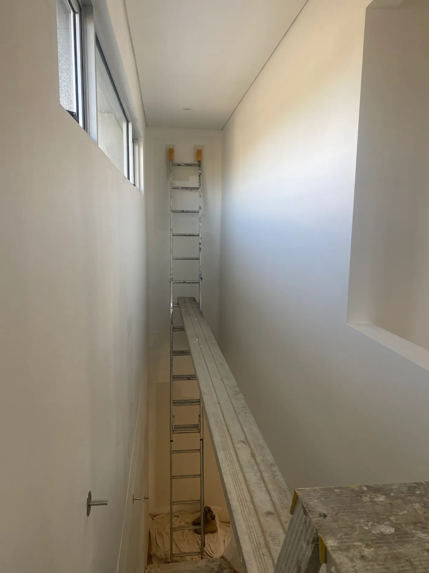 Tall, narrow internal stairwell void in a Black Rock home before painting, in bare plaster with a ladder and plank staged as a working platform