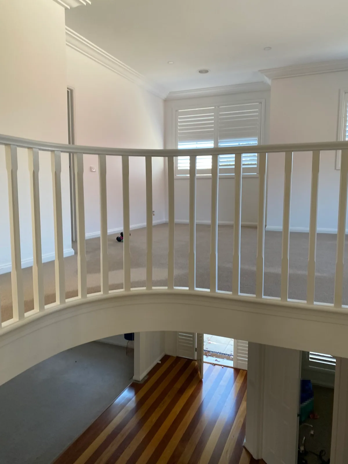 Curved white balustrade around the stair void of a Brighton home after an interior repaint, looking across the upstairs landing with white walls, cornices and shutters, above two-tone timber floorboards in the entry hall below
