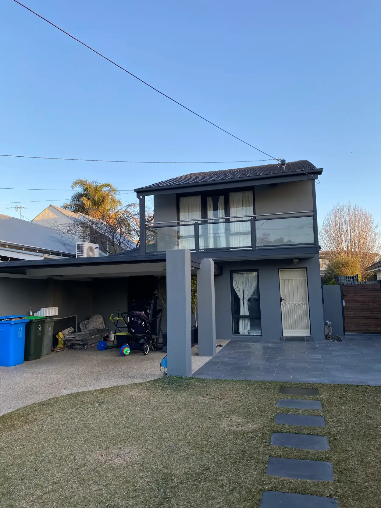 Two-storey rendered Brighton East home after exterior painting in mid grey, with charcoal fascia, window frames and balcony edge, rendered carport pillars in the facade colour and a cream security door