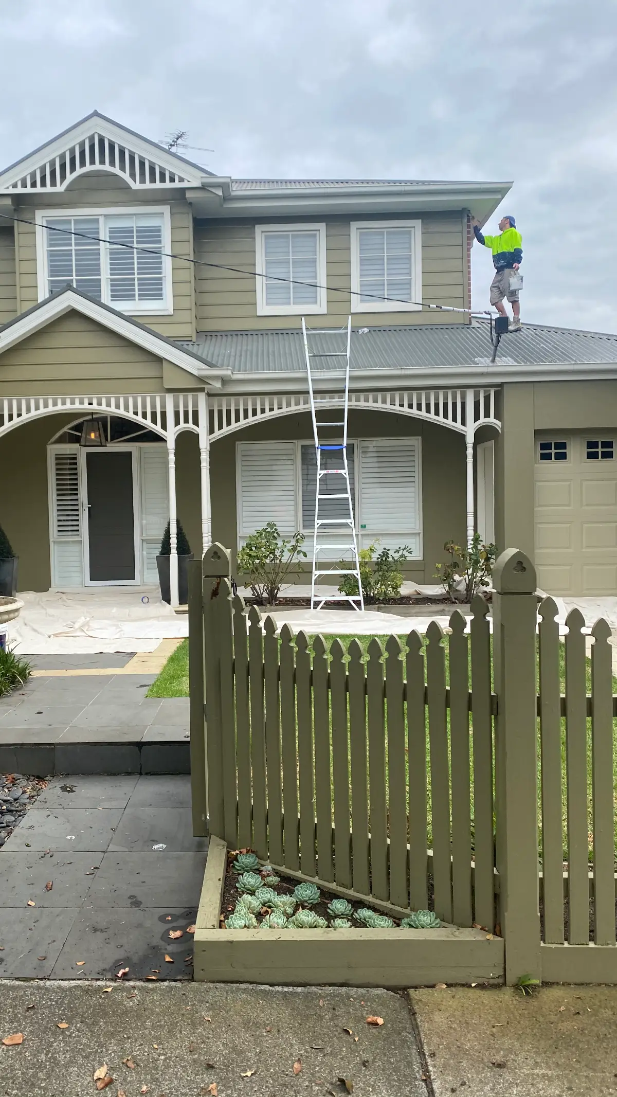 Weatherboard Brighton home in olive and cream before repainting, with a painter working the upper storey from the verandah roof
