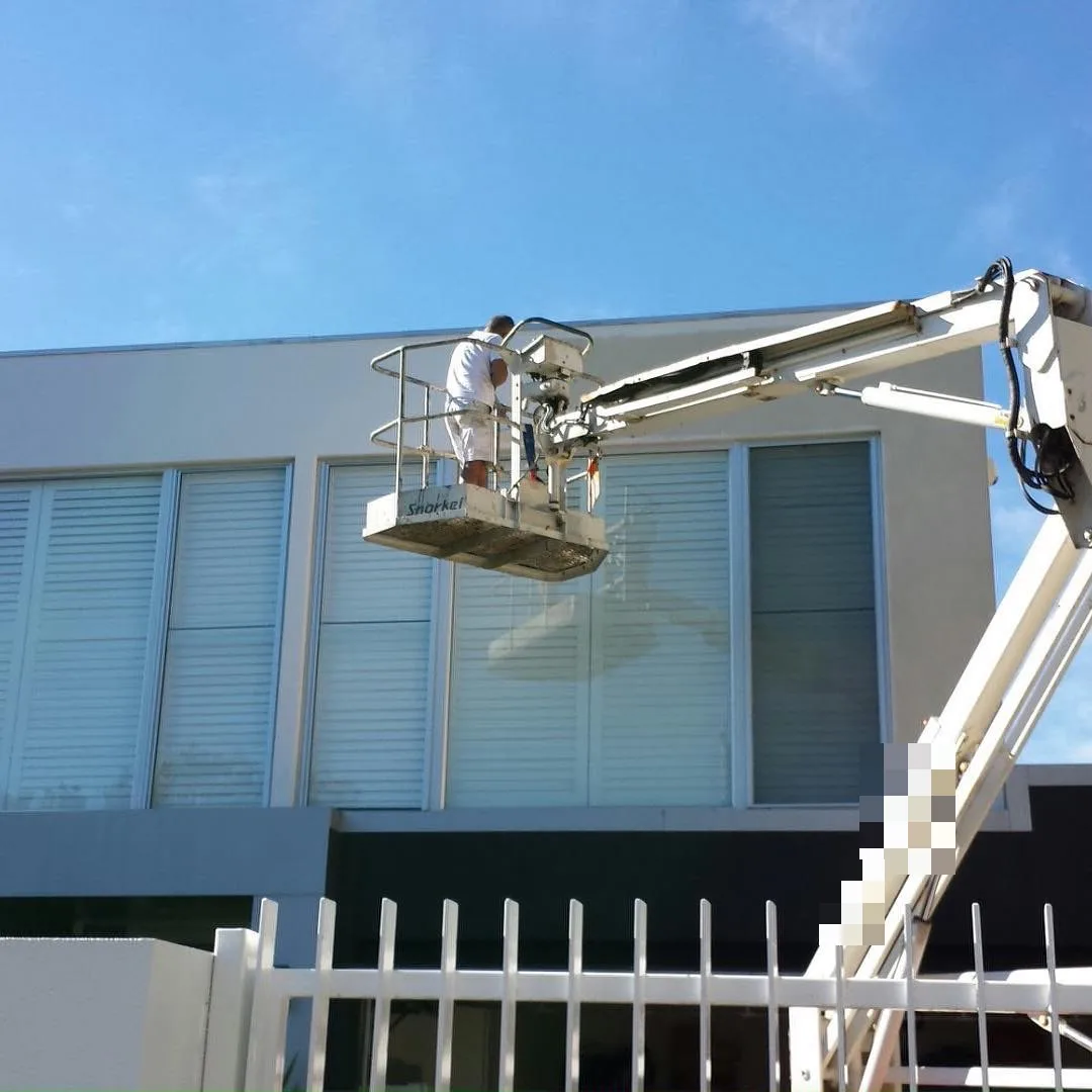 A painter in a boom lift basket working on the upper storey of a rendered two-storey home in Brighton, white render and window frames above a dark lower level