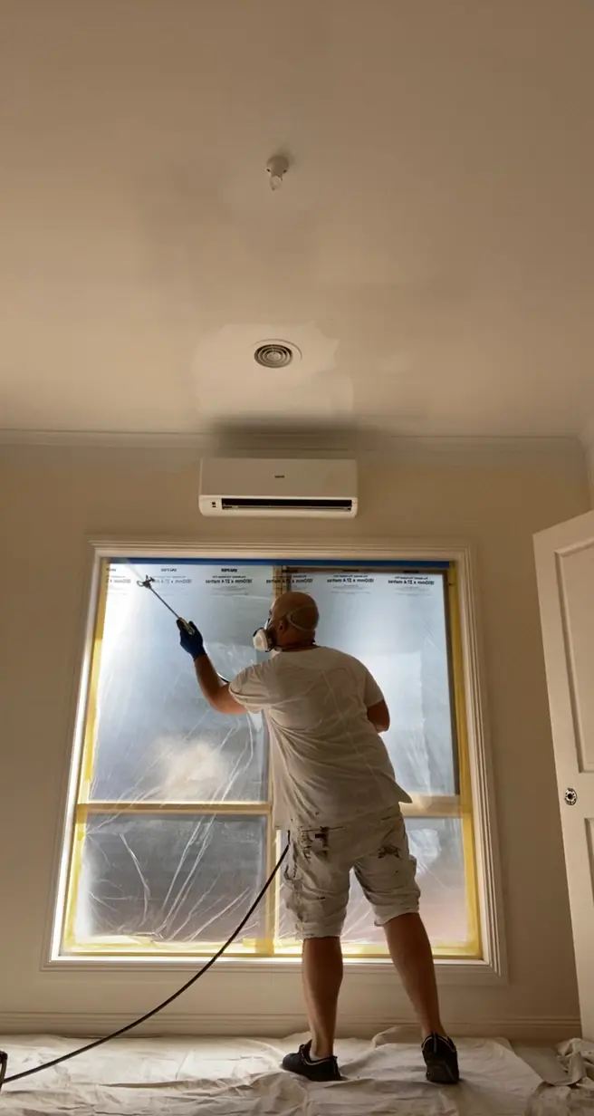Bedroom in a Brighton home mid-repaint, a Jetblack painter in a respirator spraying the frame of a large window that is masked in plastic, with the floor sheeted
