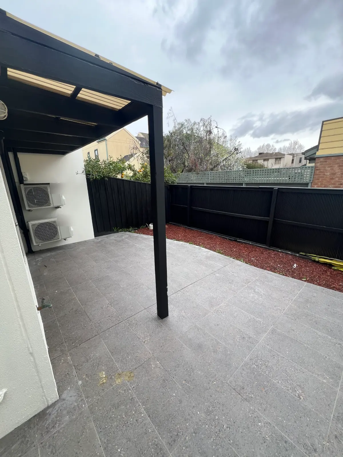 Rear courtyard of a Carlton townhouse seen from beside the house, with a black timber pergola post and beams against a black fence line