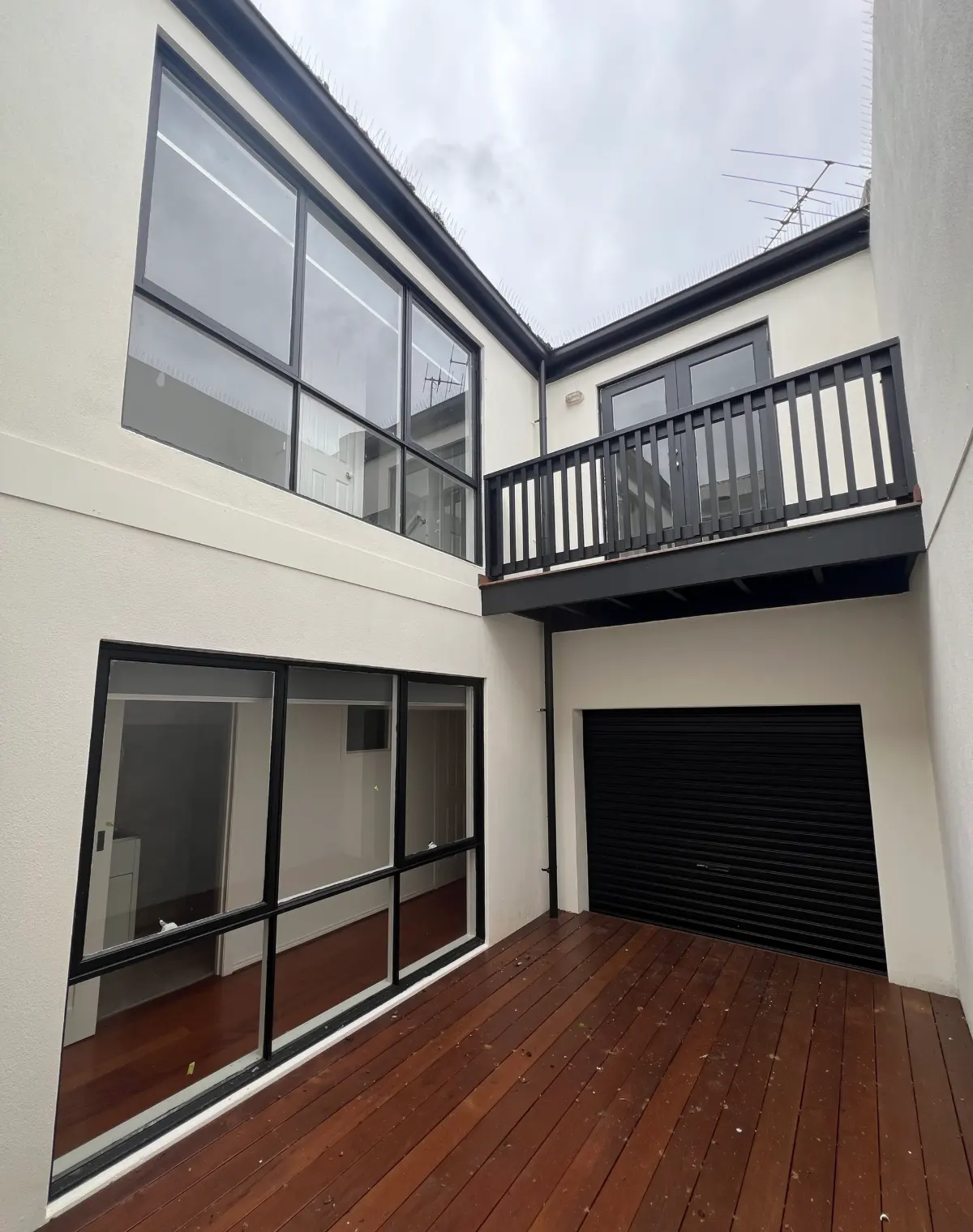 Enclosed internal courtyard of a Carlton townhouse with white rendered walls, large black-framed windows, a dark upper balcony and timber decking