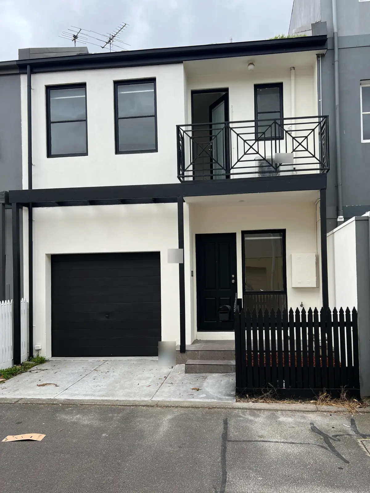 Lane frontage of a two-storey rendered townhouse in Carlton after an exterior repaint, with white render, black window frames, a black geometric balcony balustrade, black pergola beams, a black front door and a black picket fence