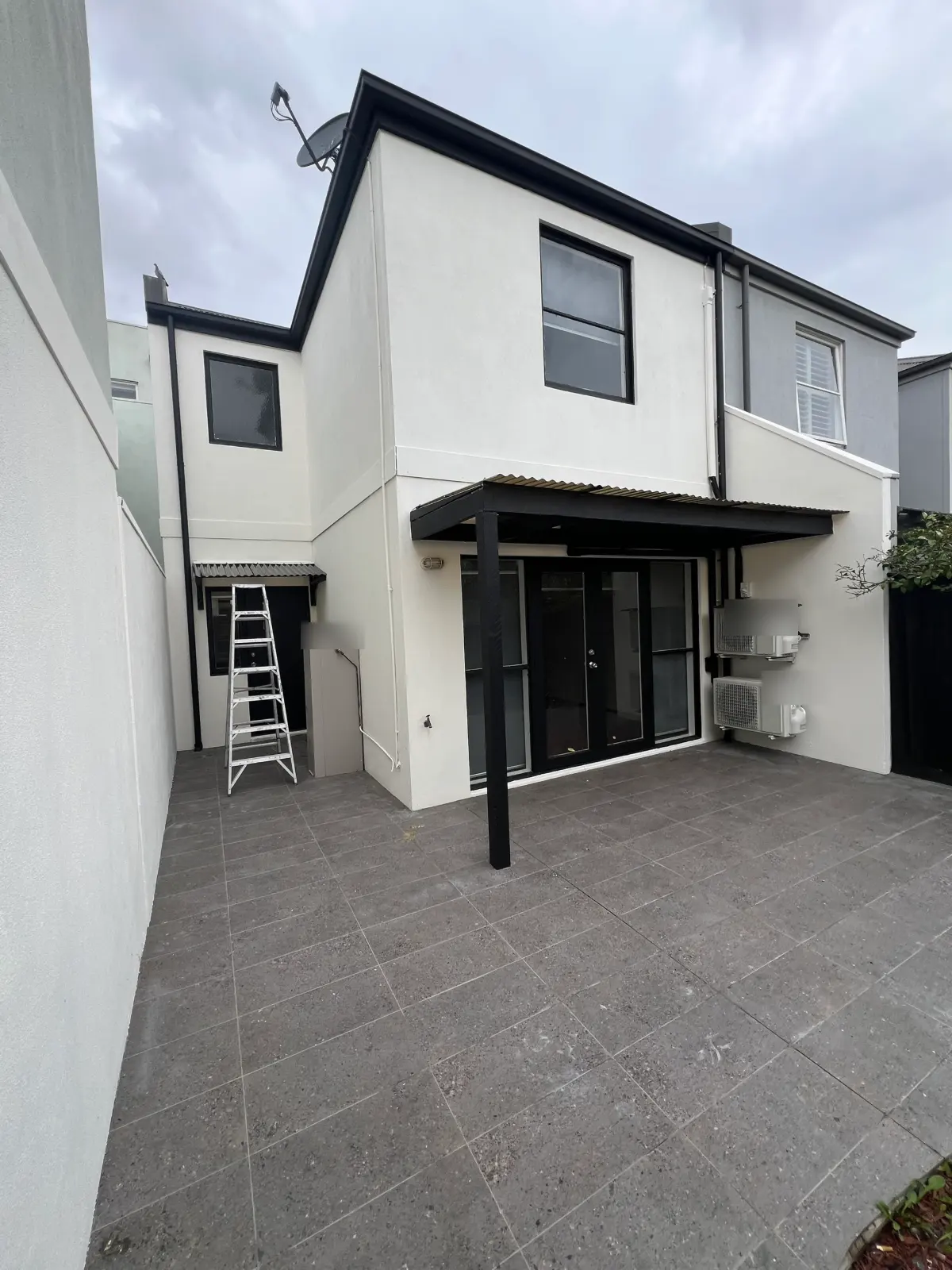 Rear elevation of a rendered Carlton townhouse in white with black gutters, downpipes, window frames and a black timber pergola over a tiled courtyard