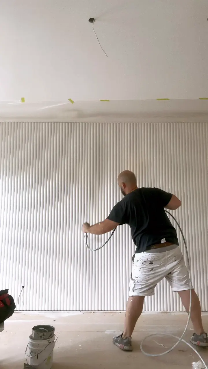 A Jetblack painter spraying a white vertical-groove (VJ) panelled wall in a Caulfield home, with the ceiling line masked and a paint tin on the sheeted floor