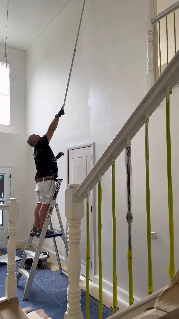 Tall stairwell in a Chadstone home mid-repaint, a Jetblack painter on a stepladder rolling the upper wall with an extension pole, the turned stair balusters wrapped in tape