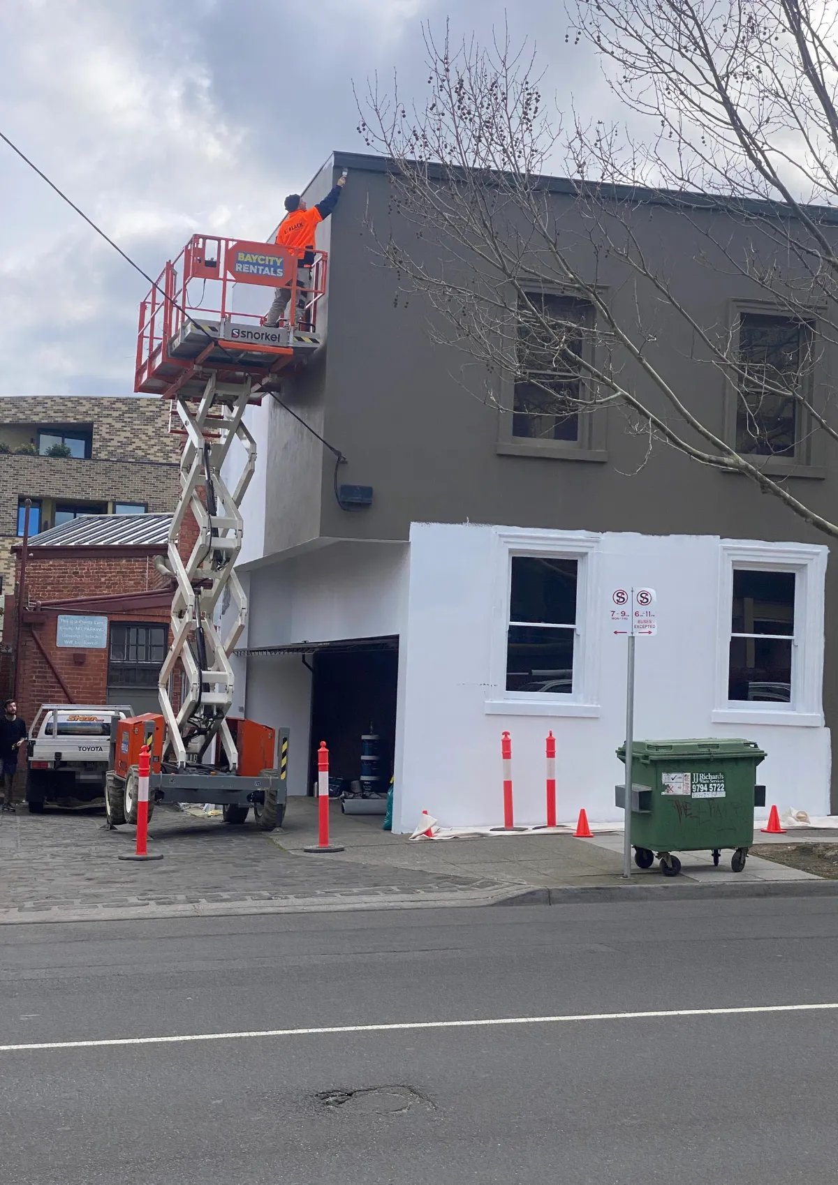 Jetblack painter cutting in the top of a two-storey commercial facade from a scissor lift, with cones and pedestrian management set out across the footpath below