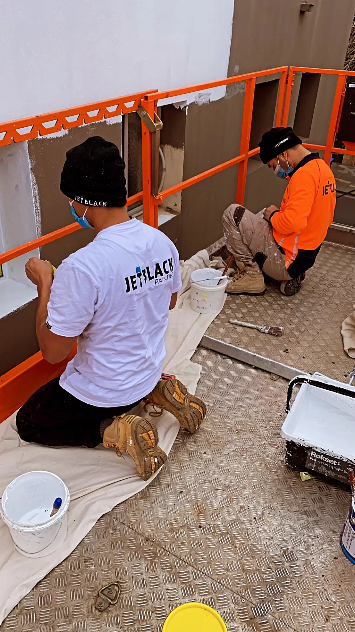 Two Jetblack painters in branded workwear cutting in window reveals on a rendered facade from an elevated work platform