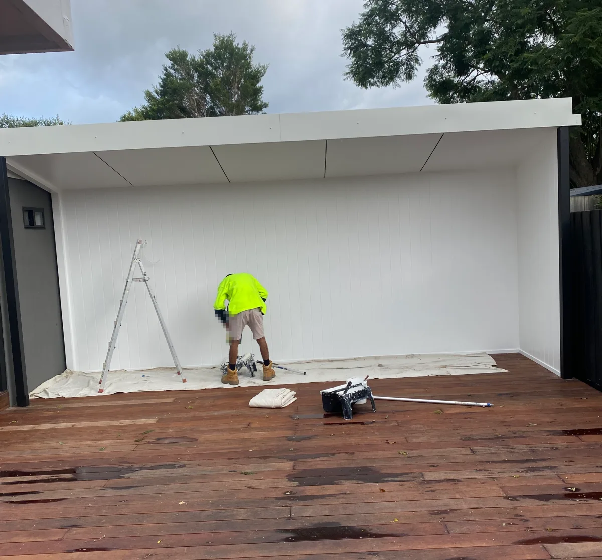 A painter in hi-vis at work in an outdoor room in Dingley Village, with white vertically grooved wall panels and a white ceiling and fascia above a timber deck, drop sheets laid along the wall