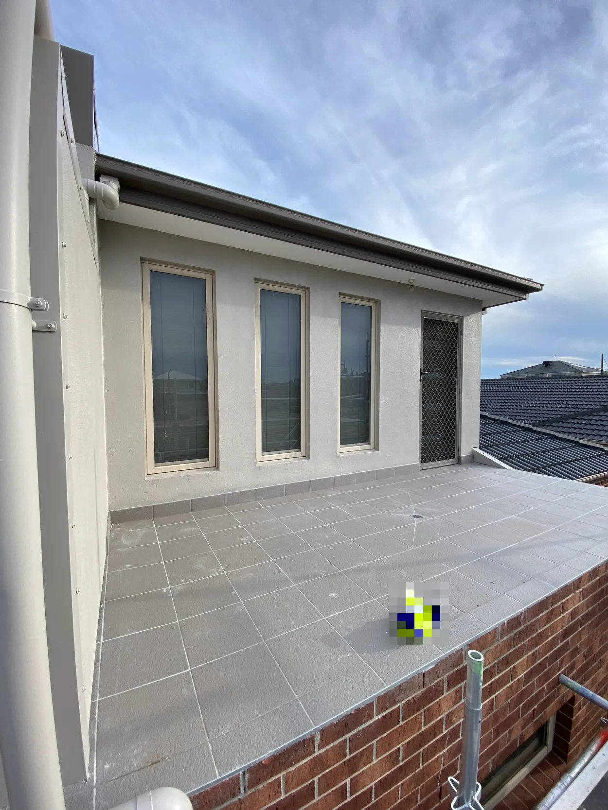 Upper-storey rendered wall of a Doncaster home with three tall windows and a screen door opening onto a tiled roof deck, with scaffold at the deck edge above the brick ground floor