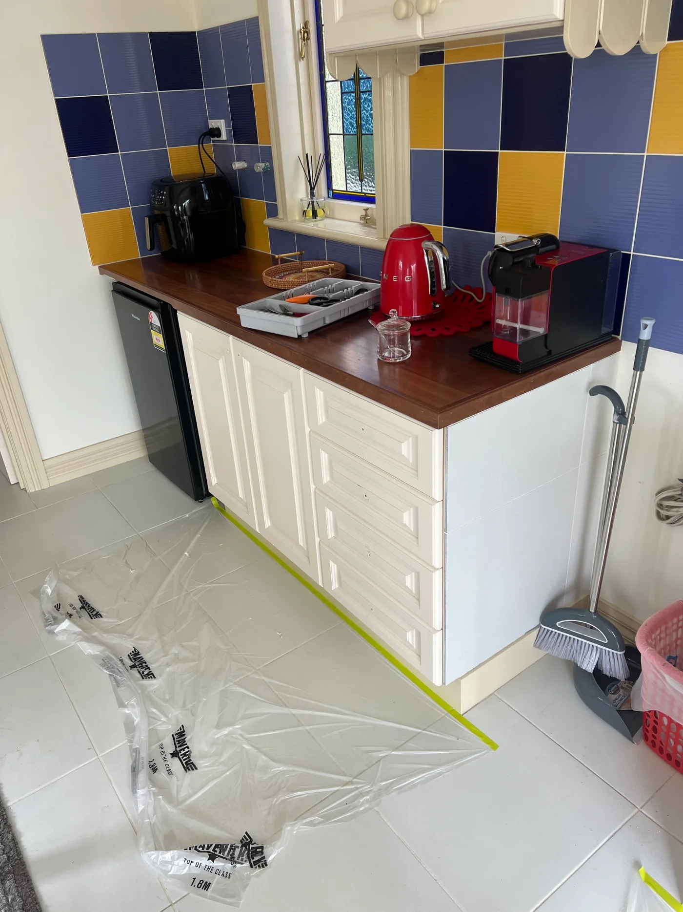 Kitchen in a Dromana home before cabinet resurfacing, with a retro blue and gold tile splashback, timber benchtop and white cabinetry, floor sheeted for protection