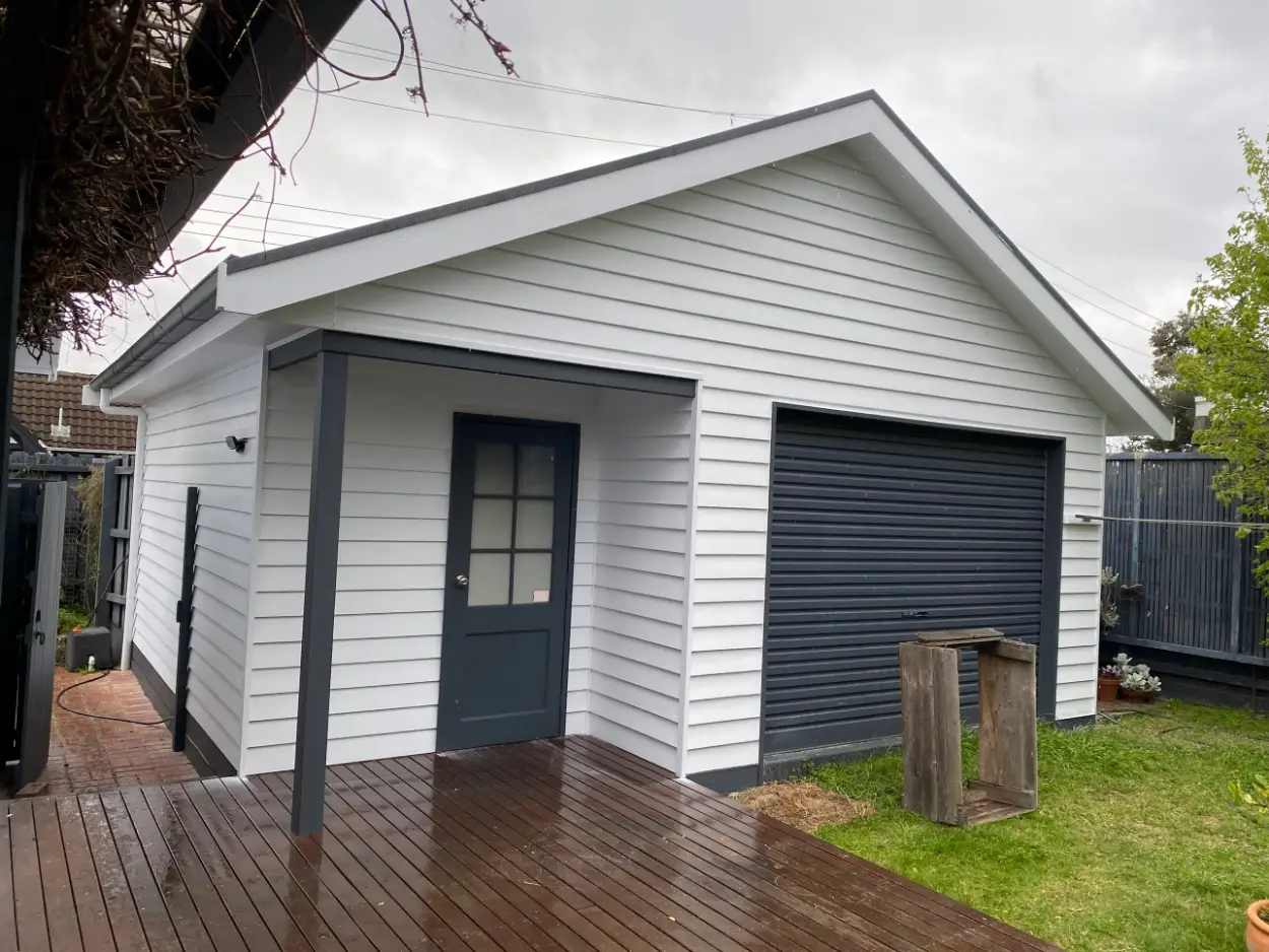 Detached weatherboard garage and studio at an Edithvale property after painting, finished in white with charcoal posts, door and roller door beside a timber deck