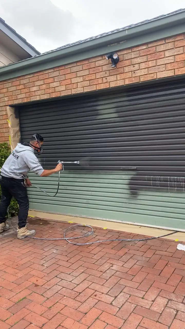 A Jetblack painter in a respirator spraying a green garage roller door charcoal, with the brickwork either side masked
