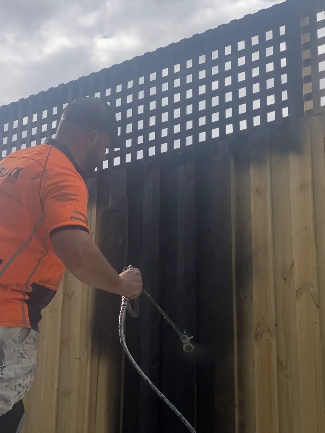 A Jetblack painter spraying a new timber paling fence and lattice topper black, with the first boards already coated