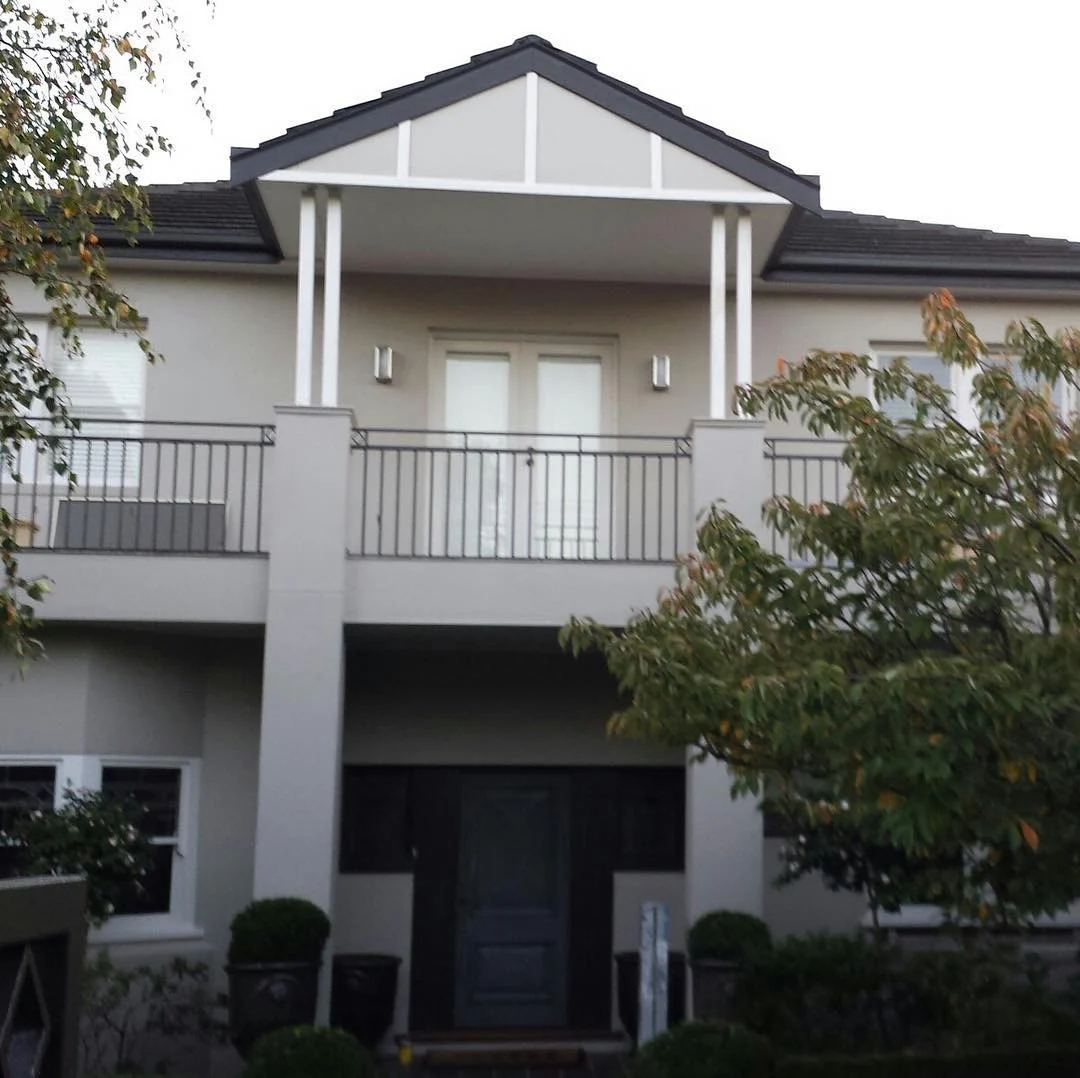 Two-storey rendered Hampton home after an exterior repaint, greige render with white posts and gable battens, a charcoal roofline and a black iron balcony balustrade