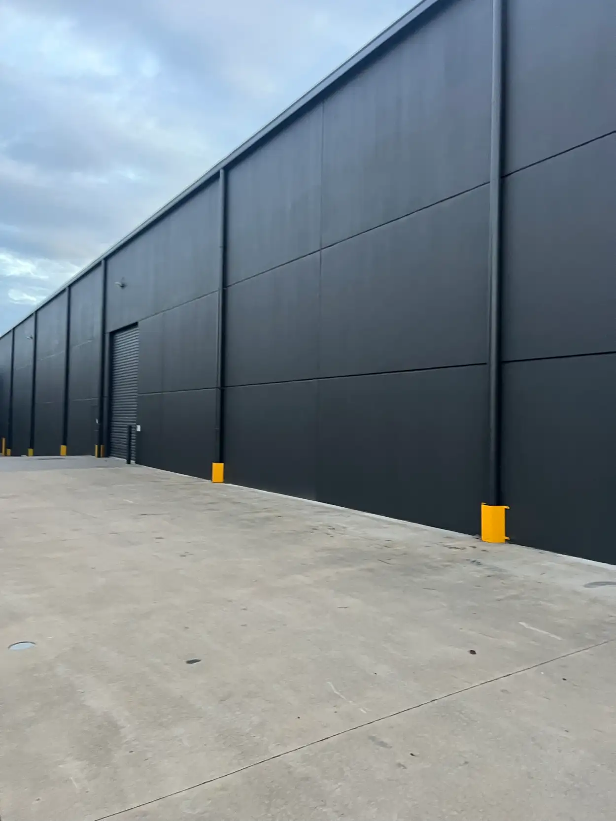 Long charcoal side wall of a Heatherton warehouse after exterior painting, with matching downpipes and yellow bollard guards at ground level
