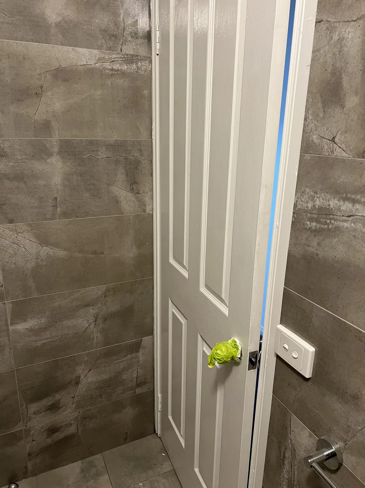 Four-panel bathroom door freshly painted white between grey tiled walls, with the handle wrapped in masking tape