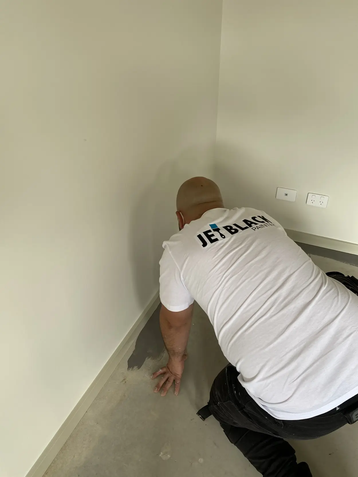 A Jetblack painter kneeling to cut in along the skirting of a freshly painted room with a concrete floor