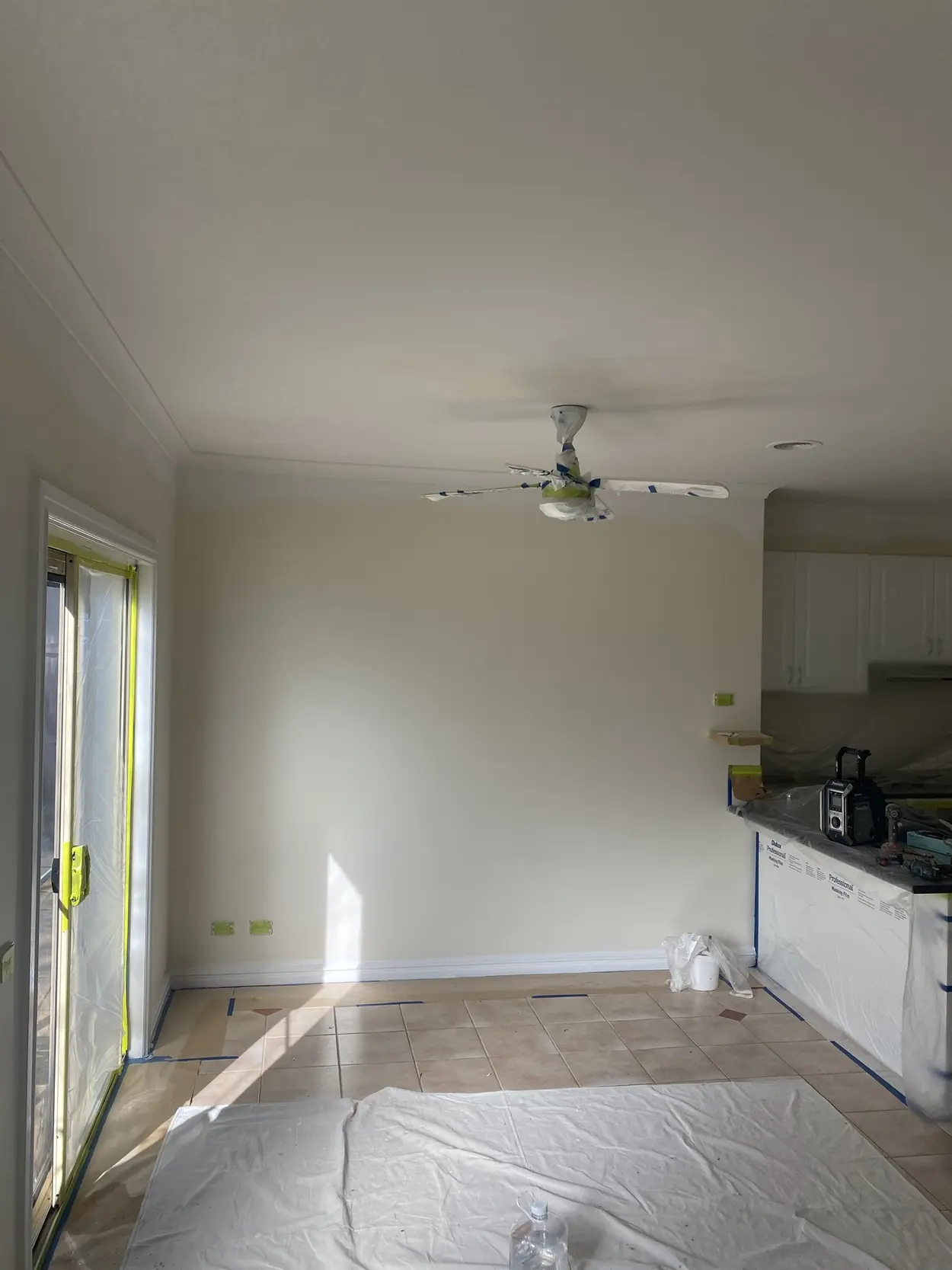 Dining area mid-repaint, with the ceiling fan wrapped in plastic, the sliding door and kitchen bench masked, and the tiled floor covered