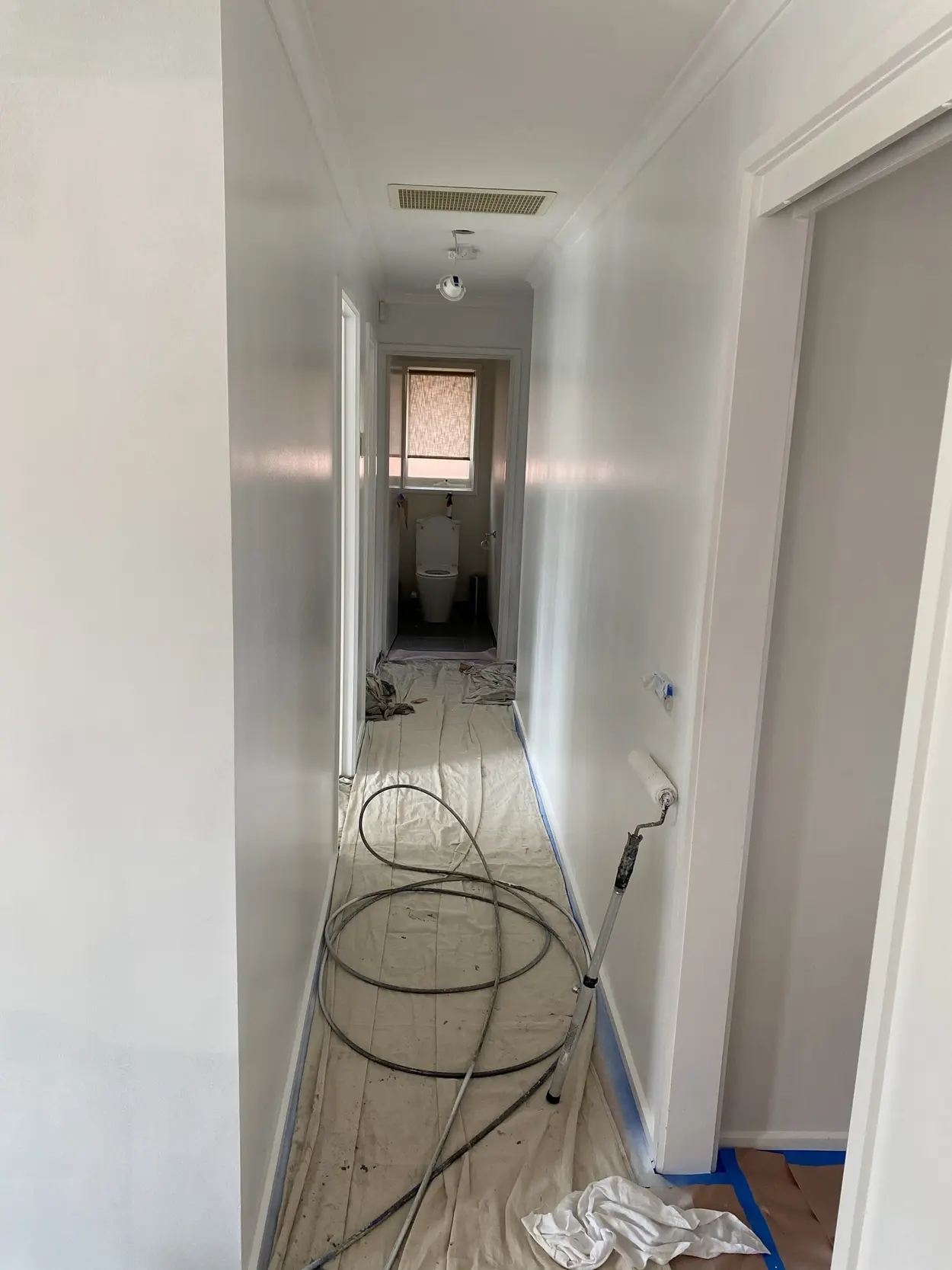 Long hallway mid-repaint, with the floor sheeted and the skirting taped, an airless spray hose coiled on the drop sheets and a roller on a pole