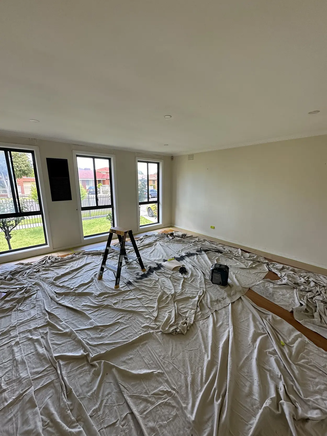 The same living room from the other side, sheeted wall to wall, with cream walls and a row of black-framed windows onto the front garden