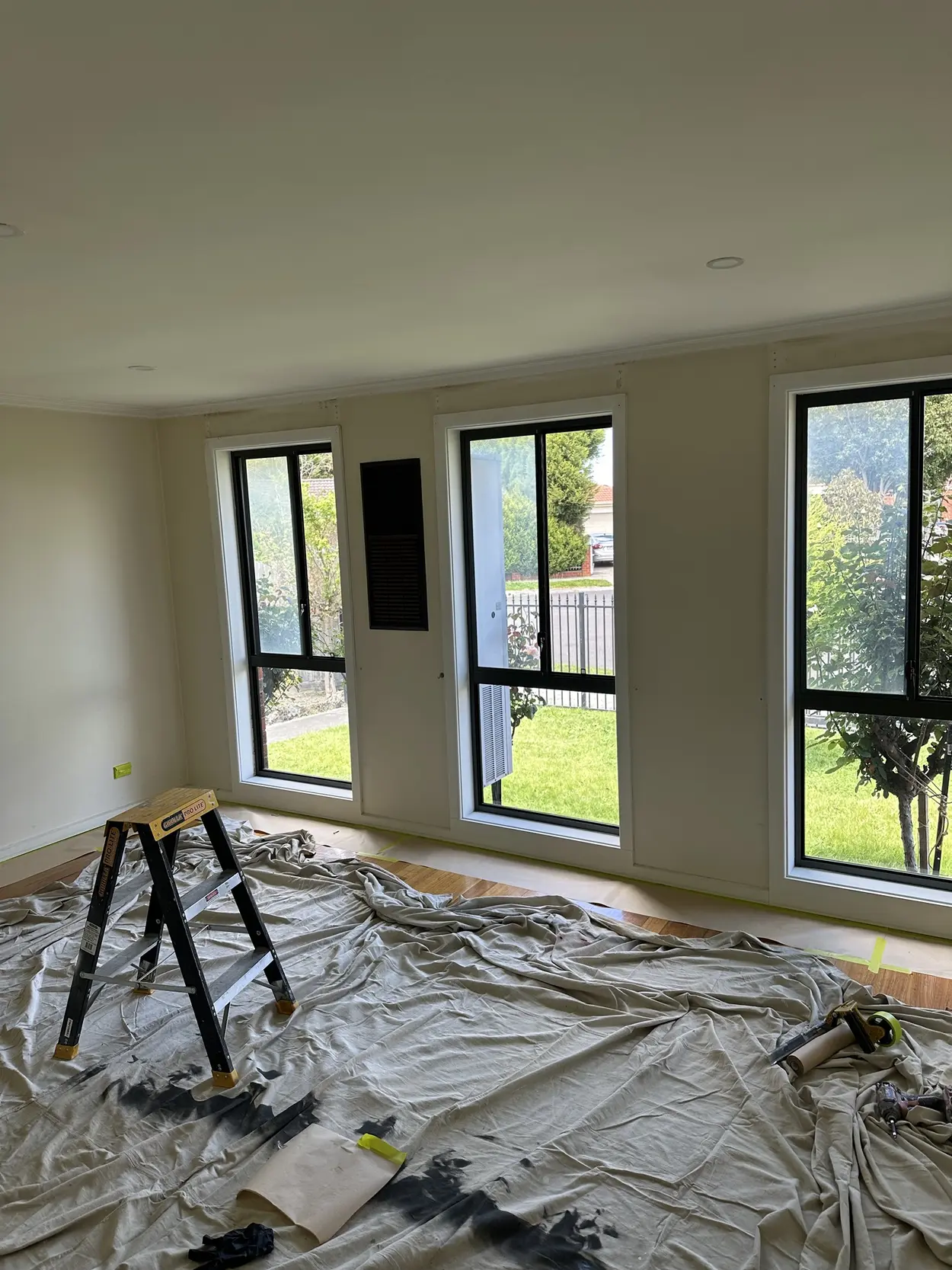 Living room during an interior repaint, with three tall black-framed windows in white architraves, drop sheets over the floor and a stepladder