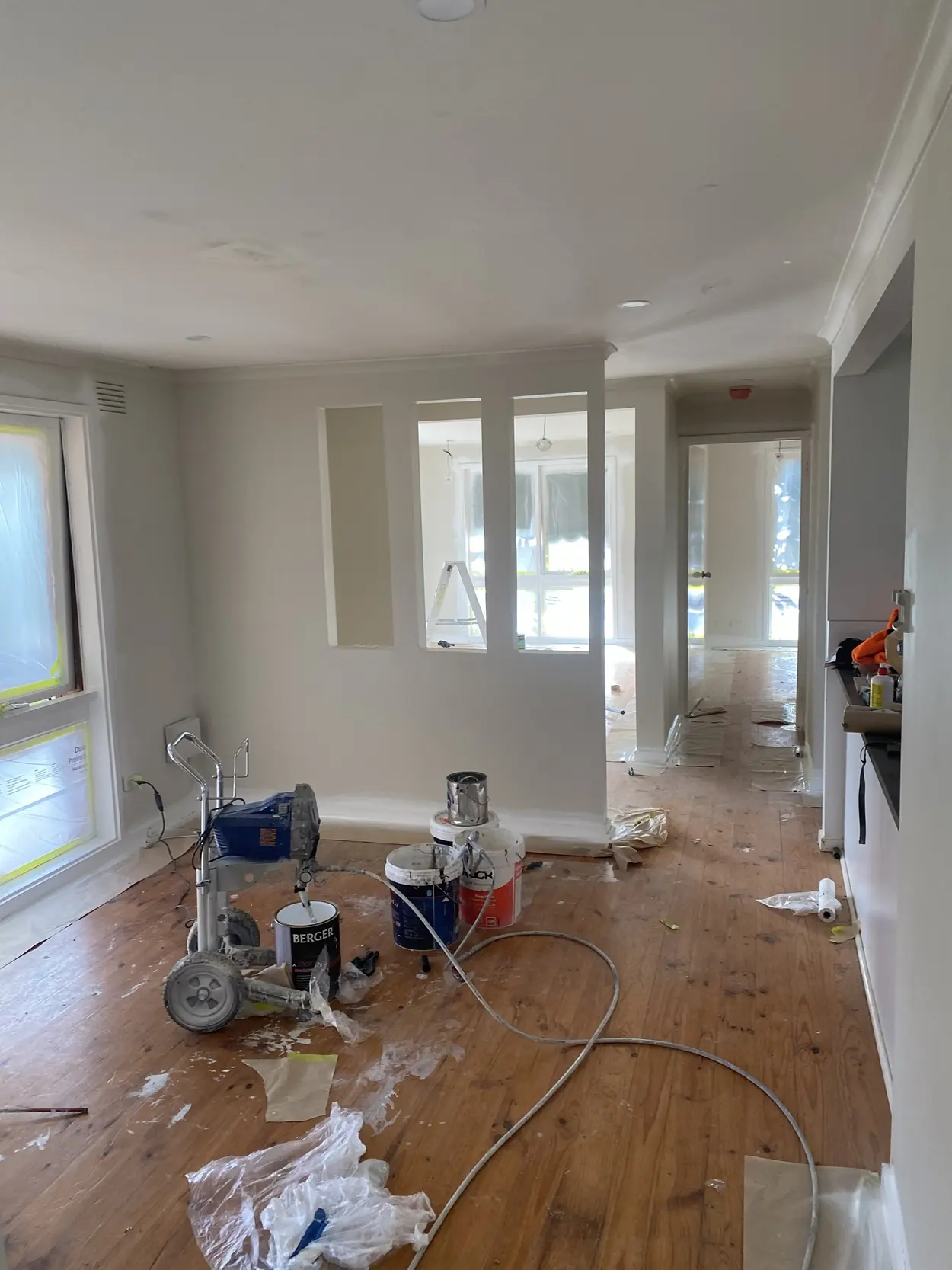 Open-plan room mid-repaint, with an airless spray rig and paint tins on the floorboards, masked windows and a feature wall with three openings