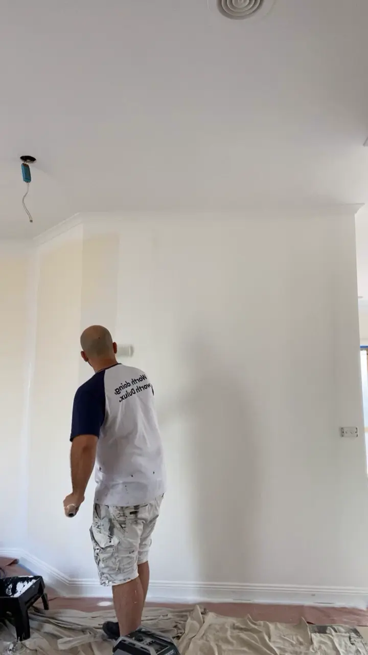 A Jetblack painter rolling a tall white living room wall with an extension pole, the floor sheeted
