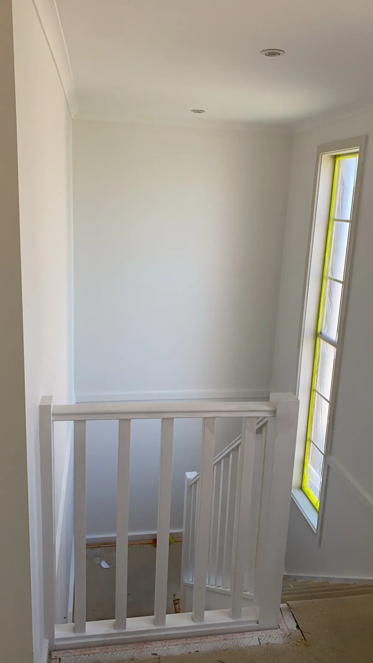 Stairwell after an interior repaint, with a white timber balustrade and a tall narrow window taped and masked beside the stairs