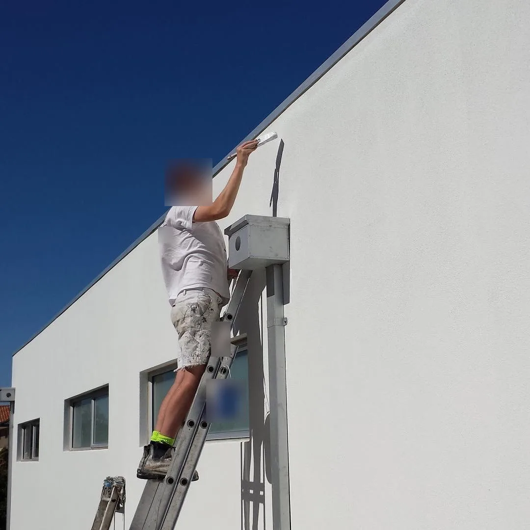 A painter on an extension ladder working along the top edge of a white rendered wall on a Malvern East property, below a metal parapet capping against a clear blue sky