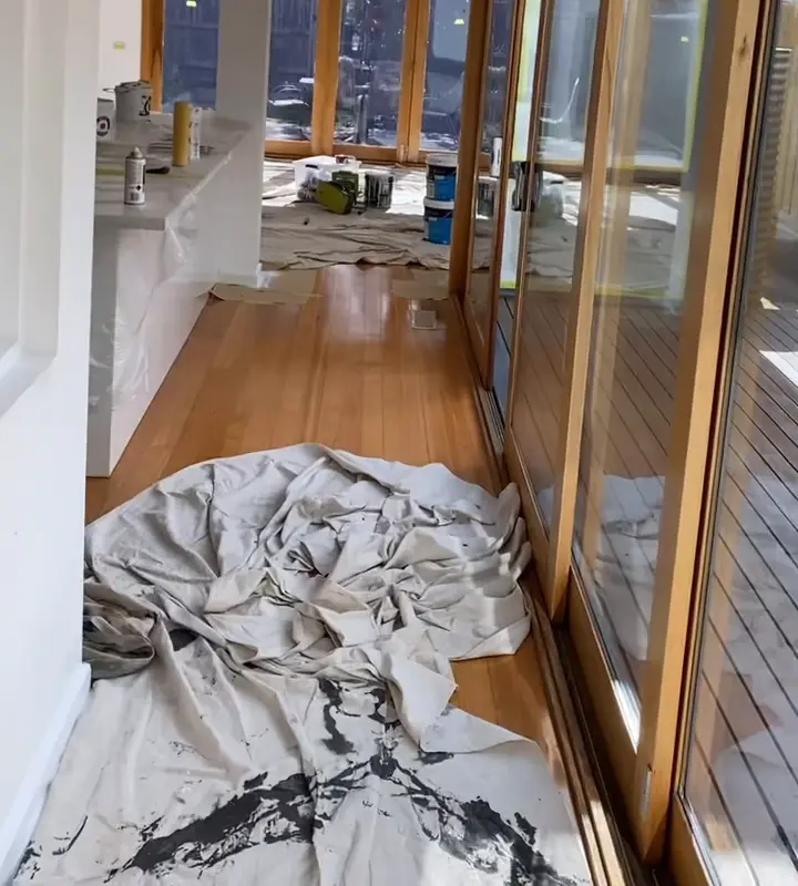 Open-plan living area in a McKinnon home during an interior repaint, with drop sheets on the timber floor, the kitchen bench wrapped in plastic and a row of timber-framed sliding doors