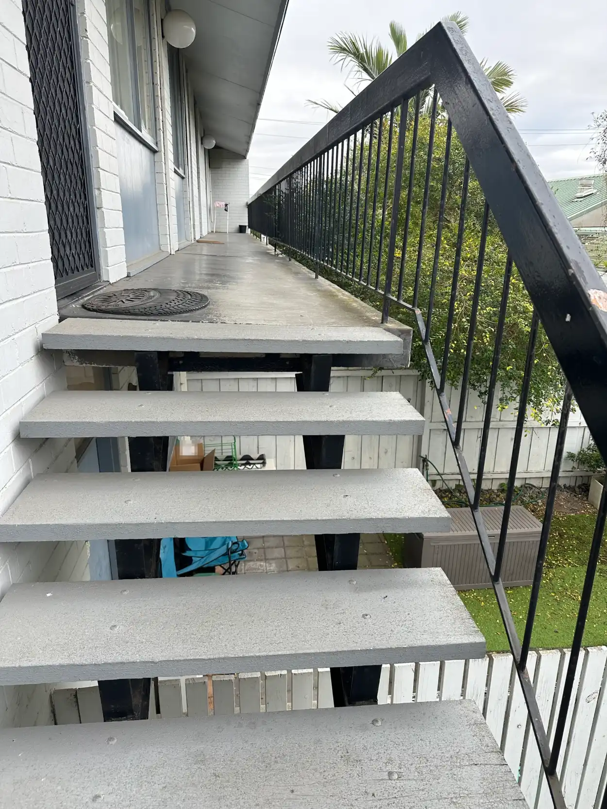 Upper landing and walkway of a Mentone apartment building before repainting, with pale, scuffed stair treads and rust showing through the coating on the black steel handrail