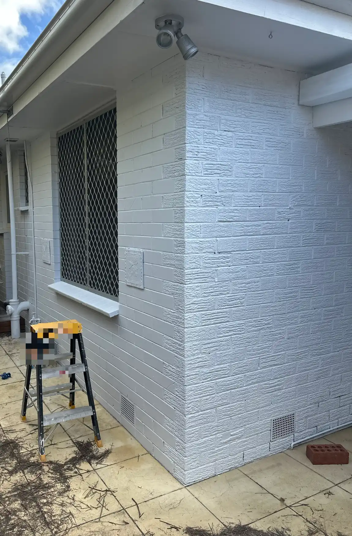 Corner of a single-storey brick home in Mentone after painting, with rough-faced brick on the corner, smoother brick along the side wall, white eaves and gutter, a window with a security screen, and a stepladder on the paving