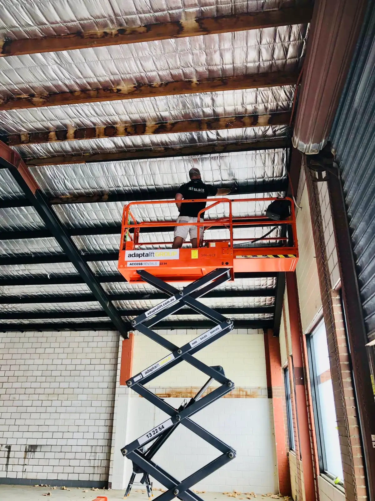 Interior of the same Mordialloc warehouse during painting, with a painter on a scissor lift working at the insulated roof structure above painted brick walls