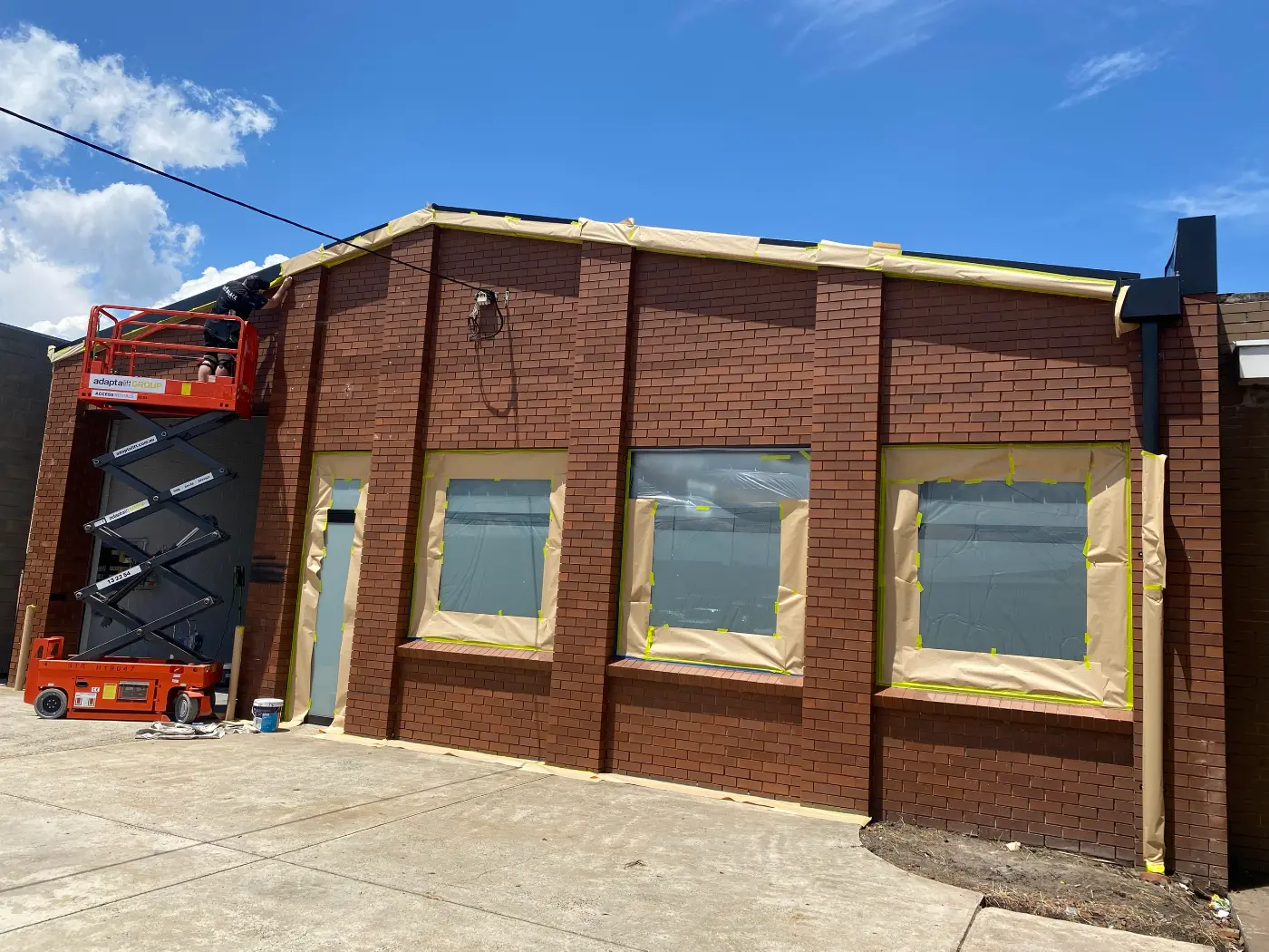 The same Mordialloc commercial facade mid-job, with masking paper and tape over the roofline and every window and a painter working from a scissor lift